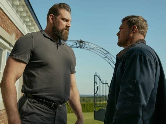 Olivier Richters as Paulie and Alan Ritchson as Jack Reacher in "Reacher." The two men are staring at each other as they stand outside a building, and fields of green grass can be seen in the background.