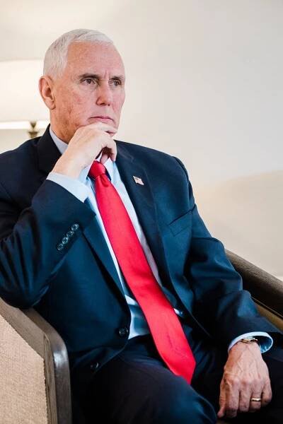 Former Vice President Mike Pence sitting in a tan chair wearing a navy blue suit with red tie and placing his finger on his chin.