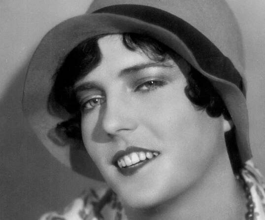 Grayscale Portrait Photo of the actress Pepi Lederer, wearing a white dress, pearl necklace and a hat, smiling