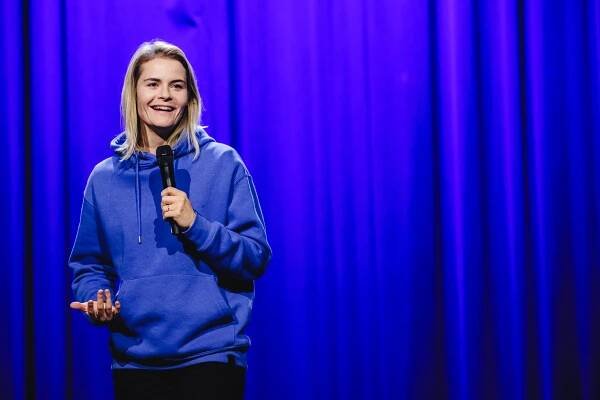 Die Schweizer Comedian Hazel Brugger in einem blauen Hoodie hält ein Mikrofon und spricht auf einer Bühne vor einem lila Vorhang.