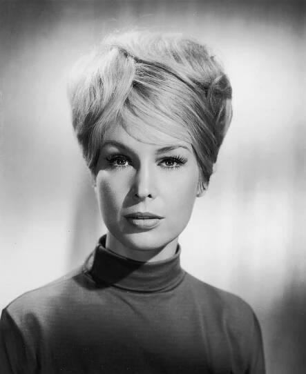 Person with short, styled hair wearing a high-collared top, looking directly at the camera in a studio portrait