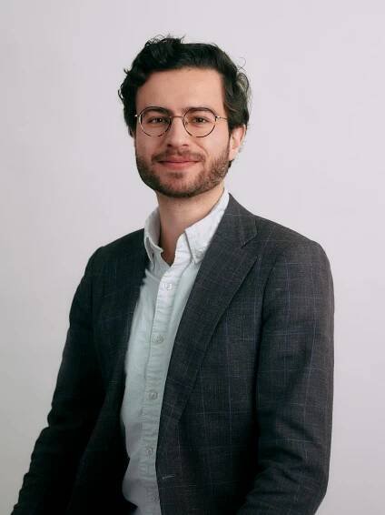 Jérôme Nicolaï, a man with glasses and a beard, wears a suit jacket and a light shirt, smiling warmly against a plain background.