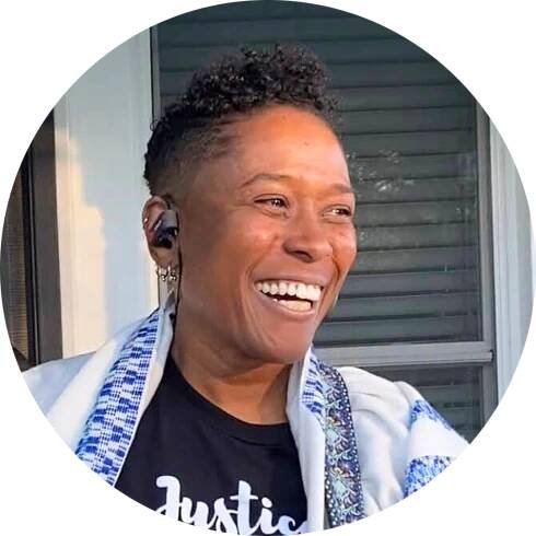 Rabbi Sandra Lawson smiling warmly, wearing a black t-shirt and headphones, standing outdoors near a window.