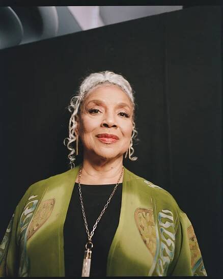 Phylicia Rashad in a green silk cardigan, black shirt and long gold necklace looks into the camera.