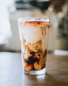 A pint-style glass sits on a brightly lit countertop, filled with cold brew coffee and ice cubes, with creamer swirling throughout as if it were just poured into the glass