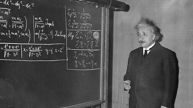 A black and white photograph of Albert Einstein in front of a blackboard