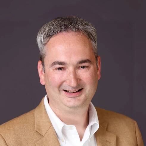 A man in a tan suit and white shirt is smiling for the camera.