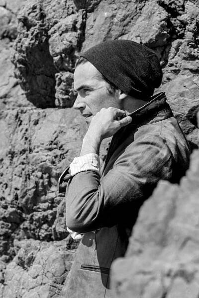 Black and white shot of Ian Harding, a man standing on a rocky wall, wearing a beanie and a grayish jacket