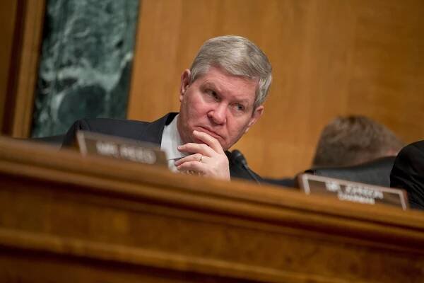 Tim Johnson, a gray-haired man with a serious expression on his face, sits at a table and strokes his chin with his left hand.