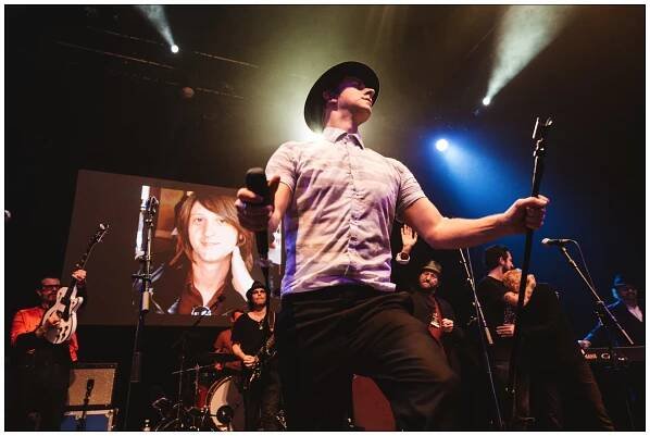 Paul smith on stage in front of a picture of Nick Alexander, Jesse Hughes, Frank Turner and Chas'n'Dave at the Shepherd's Bush Empire