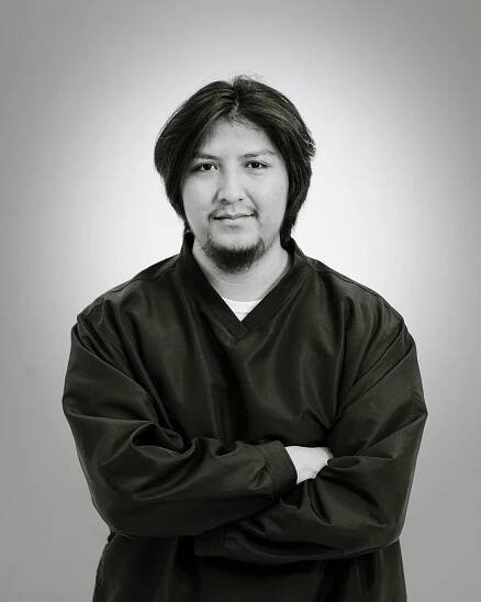 Black and white studio portrait of a person in dark v-neck top with arms crossed against a gradient background.