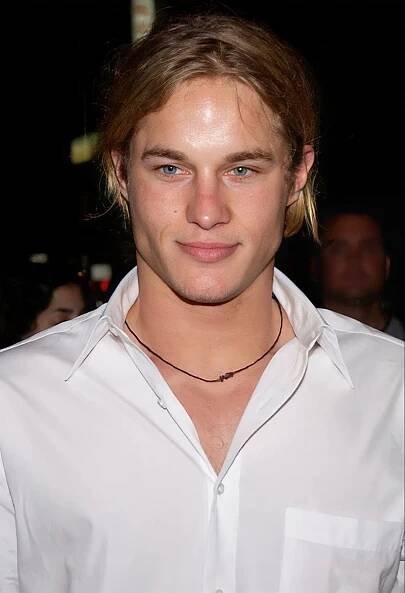 a man with long hair wearing a white shirt and looking at the camera while standing in front of a crowd