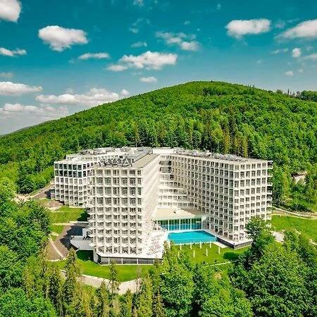 Crystal Mountain Hotel Visla Exteriér fotografie The photo shows a large, modern hotel complex nestled in a lush, green, hilly landscape under a partly cloudy sky. The hotel consists of several white, multi-story buildings with numerous windows, arranged in a somewhat sprawling layout. 

In the cen