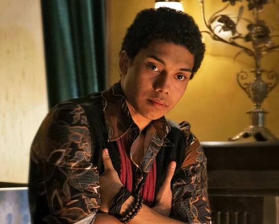 a photo of Ambrose Spellman (Chance Perdomo), a young black man, with his hands tucked up under his arms, staring intently at a character off-screen. He is wearing an ornately patternd black and bronze shirt, a waistcoast, and wooden bracelets. In the background, a living room with a yellow wall, curtains, a candelabra and a teacup.