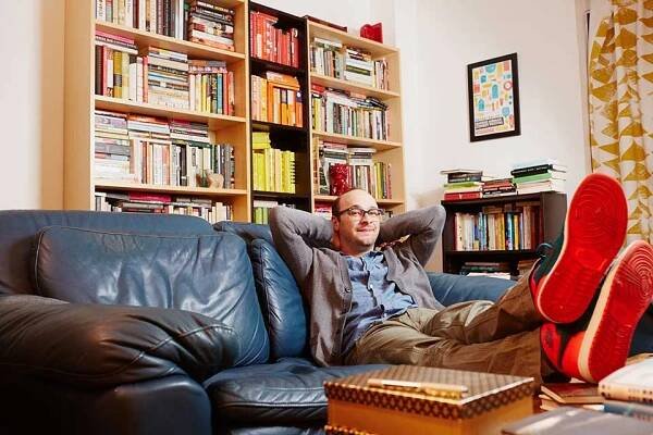 Josh Gondelman sitting on a couch with books on bookshelves in the background. 