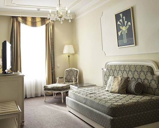 Esplanade Spa And Golf Resort Mariánské Lázně Exterior foto The photo shows a well-lit hotel room with a comfortable and elegant ambiance. The room features a large bed with a patterned headboard and a gray and gold patterned bedspread. Several pillows in varying shades of beige and gold are arranged on the b