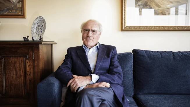 Geoffrey Palmer sits on a couch and looks at the camera.