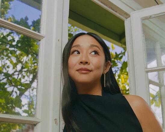 Hsu leans against the white wood frame of a space with trees and blue sky behind her. 