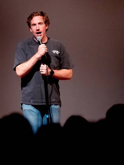 A man in a black T-shirt and jeans stands  on a stage holding a microphone to his mouth. 