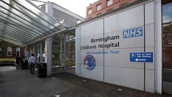 The Birmingham Children's Hospital trust sign is visible by the entrance to the hospital with glass roofing and frontage. It is black and blue writing, with a circular picture with an animated child in its centre.