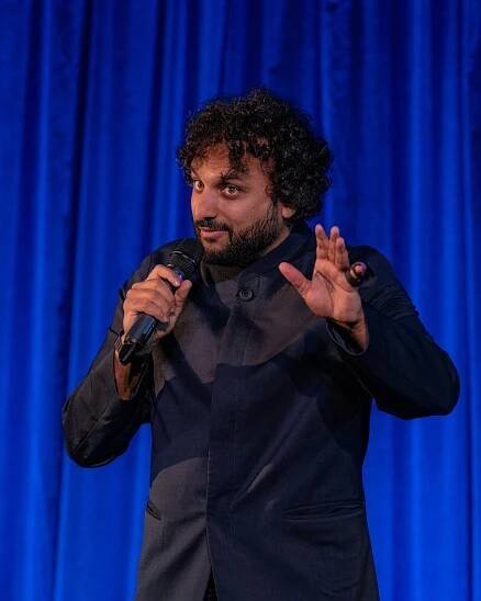 A man in a dark sports jacket holds a microphone in his right hand while standing in front of a blue curtain.