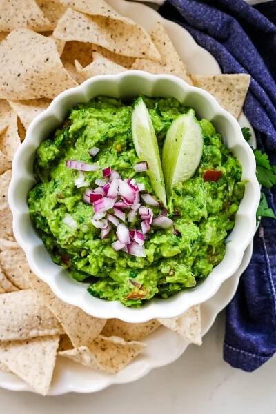 Chunky guacamole in a white bowl, with tortilla chips beside it on a white plate.