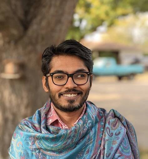 Efad is seen from the mid-torso up smiling into the camera. Efad is wearing black glasses and is draped in a soft-looking colorful blue fabric. In the background is a blurred tree and a warm-toned landscape.