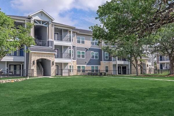 Modern apartment community featuring multiple buildings with a combination of blue and beige siding. The structures include balconies and are surrounded by well-maintained green lawns and trees. A pathway runs through the landscaped area, providing access to the entrances of the apartments.