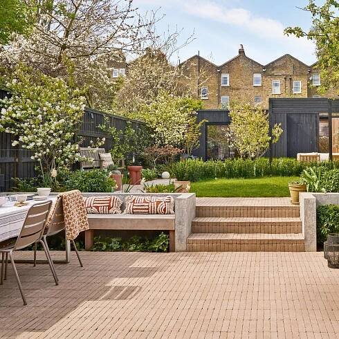a large back garden with a black time framed studio at the end, trees and lawn in the middle and a brick patio in the foreground with a dining area