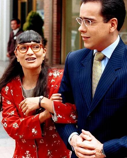 A woman in a red dress and red-rimmed glasses looks at the man in a suit next to her.