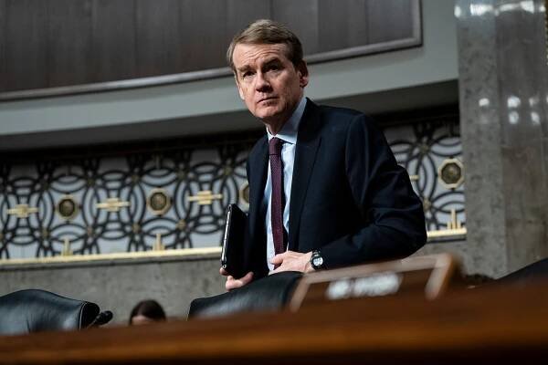 Michael Bennet, wearing a dark suit and holding a black notebook, stands behind a long desk in a Senate chamber.
