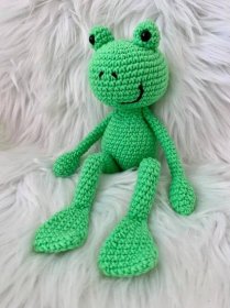 a green crocheted frog sitting on top of a white fur covered bedding