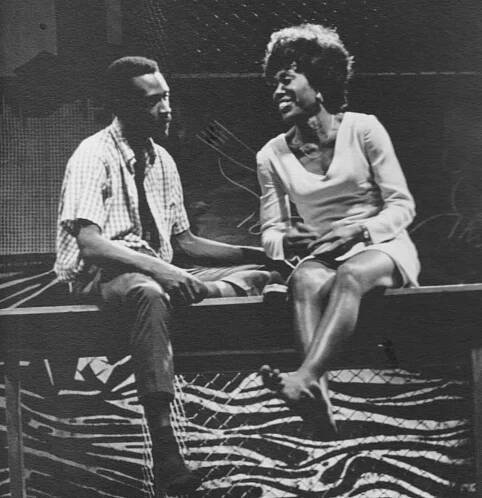 Barbara Ann Teer (right side) and Cleavon Little (left side) sit on a fence talking. Barbara's character is barefoot and there is a wall behind them decorated in tiger print.