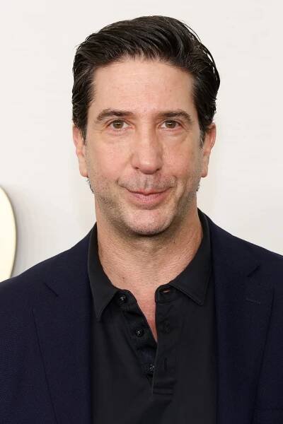 David Schwimmer in a black polo shirt and dark blazer is looking at the camera against a plain background