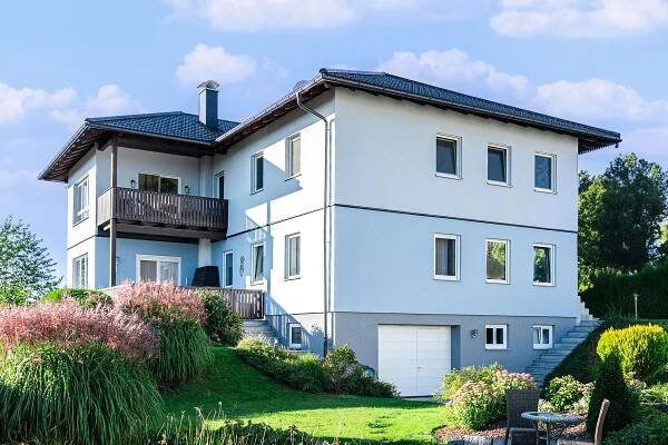 Modernes zweistöckiges Haus in Österreich mit dunkelgrauem Dach und Balkonen, von einem gepflegten Garten umgeben, symbolisiert Hausumbauten und Aufstockung für mehr Wohnraum.