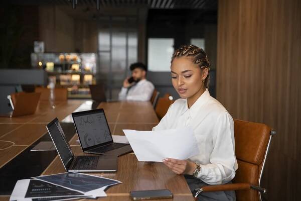 woman in a white shirt infront of two laptops looking at a paper