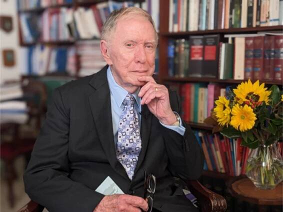 An elderly man in a suit sits in a chair, holding glasses and papers, with a thoughtful expression. Behind him are bookshelves filled with books and a vase of yellow flowers on a table.