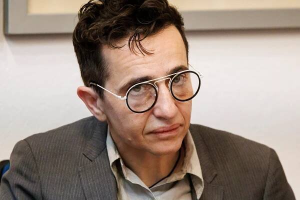 A photo  of Masha Gessen, wearing spectacles and a brown suit jacket and khaki-colored shirt.