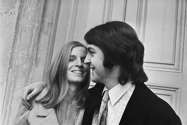 A happy straight white couple with 1960s haircuts; he is wearing a lacy shirt and has her arm around her.