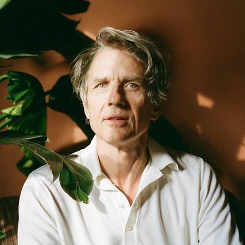 Head shot of Dean Wareham sitting in a room with a houseplant.