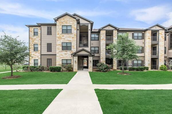 A modern apartment building with multiple stories, featuring a mixture of stone and siding exterior. There are well-maintained green lawns and small trees flanking the pathway leading to the entrance, which has a covered porch area. The sky is clear and blue, enhancing the inviting atmosphere.