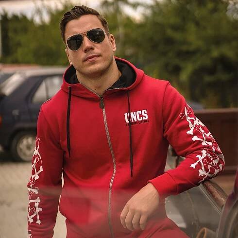 Muž v červené mikině UNCS s bílými prvky s autem a stromy v pozadí.