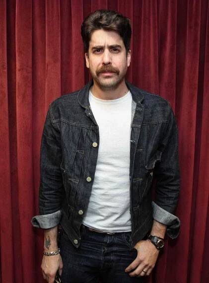 Adam Goldberg in jean jacket in front of red curtains.