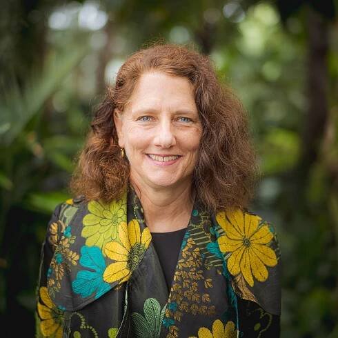 A person in a black, yellow, green, and blue blazer embroidered with flowers poses for a headshot inside a space full of green foliage