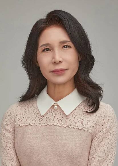 Woman with dark hair wearing a pink lace sweater and white collared shirt; neutral background.