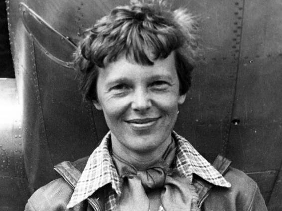 A black-and-white photo of Amelia Earhart, smiling for the camera, wearing a flight jacket. Her hair is cut short and tousled.