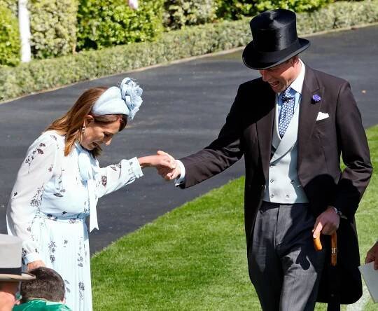 Carole Middleton wearing a blue floral dress and headband holding her hand out to Prince William, dressed in a morning suit and top hat, at Royal Ascot as they laugh