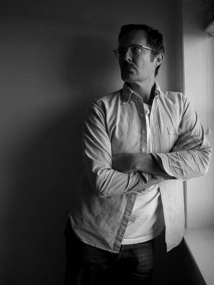 In a black and white image, a man in an unbuttoned long-sleeve shirt with a white T-shirt underneath stands indoors by a window with his arms crossed.