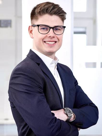 Junger Mann im Anzug mit Brille und Uhr, lächelnd mit verschränkten Armen vor einem hellen Bürohintergrund.