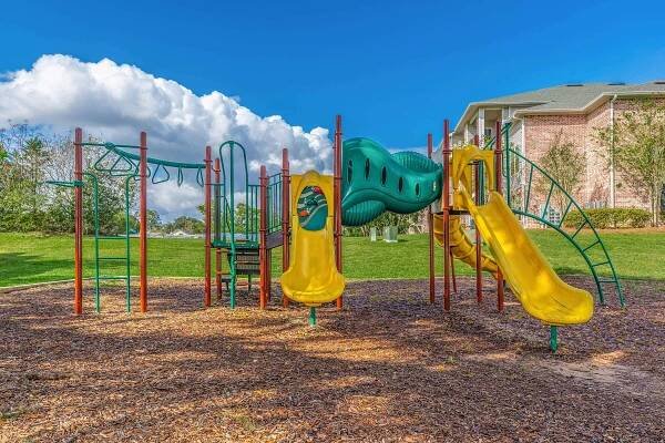 A colorful playground with green and yellow slides, climbing structures, and monkey bars, set on a sunny day under blue skies with fluffy white clouds. The playground is surrounded by grassy areas and trees, providing a fun and inviting space for children to play.
